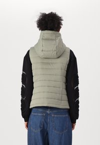 Ljust grön vadderad väst med en huva, med horisontell kviltad textur, bärs över en svart topp med intrikata detaljer och blå jeans.
