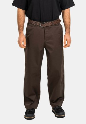 Mann trägt dunkelbraune, gerade geschnittene Hose, einen schwarzen gewebten Gürtel, schwarze Schuhe und ein schwarzes Hemd, steht vor einem schlichten Hintergrund.