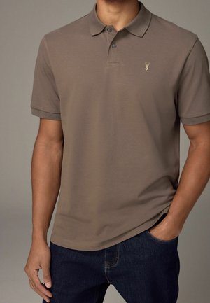 Homme portant un polo manches courtes taupe avec un petit logo de cerf sur la poitrine, associé à un jean bleu foncé, main dans la poche.