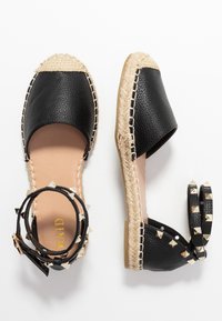 Sandalias espadrilles de cuero negro con suelas de yute y correas en el tobillo adornadas con tachuelas doradas en forma de pirámide, mostradas desde vistas superiores y laterales.