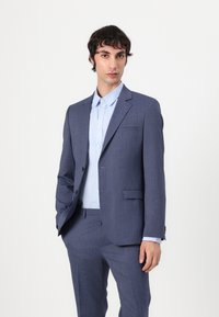 Zils pārbaudīts uzvalks ar šauru piegriezumu, vienas rindas žakete, ar izgrieztiem atlokiem un atbilstošām biksēm. Apvienots ar gaiši zilu pogājamu krekli.