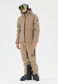 Beige isolierte Skijacke mit Reißverschlussfront, schwarzen Reißverschlusstaschen, kombiniert mit passenden beige Skihosen mit schwarzen Akzenten und Reißverschlüssen.