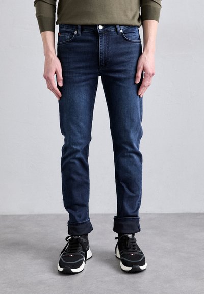 Ciemnoniebieskie jeansy o slim fit, z klasycznym pięciopocketowym wzorem i lekkim przetarciem. Noszone z czarnymi sportowymi sneakersami.