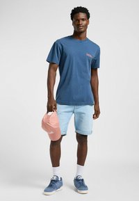 Sininen puuvillapaita, jossa on pieni vaaleanpunainen logo, yhdistettynä vaaleisiin farkkushortseihin, pidellen vaaleanpunaista baseball-lakkia, viimeisteltynä sinisillä lenkkareilla ja valkoisilla sukilla.