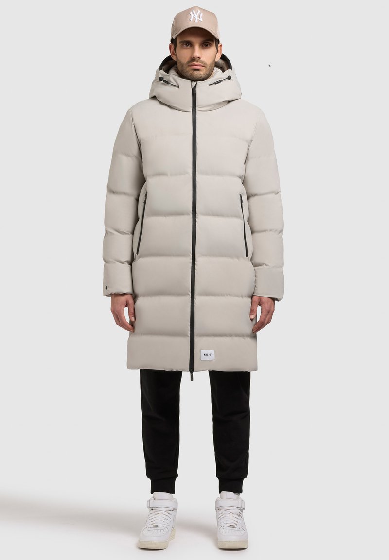 Lys grå pufferjakke med hætte, der har en frontlynlås, to sidelommer og sorte detaljer. Båret over sorte bukser og hvide sneakers.