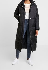 Lång svart pufferjacka med hög krage och dragkedja, bärs över en vit mönstrad tröja och denimjeans, i kombination med vita sneakers.