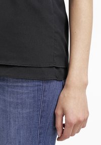 Personne portant un haut noir superposé et un jean bleu, avec le bras droit détendu le long de son corps contre un fond blanc.