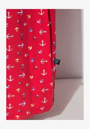 Tela roja con un patrón de pequeños anclajes multicolores en blanco, negro, azul y amarillo, con una etiqueta de ropa negra en la costura lateral.