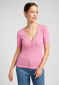 Lyserød ribbet kortærmet top med en dyb V-udskæring og tre knapper foran. Blød tekstur med en tætsiddende pasform, parret med blå jeans.