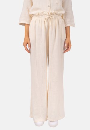Frau trägt lockere beige Textur-weite Hose mit Kordelzug in der Taille und passendem Hemd, steht mit entspannten Händen an den Seiten.