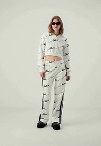 Woman wearing white crop sweatshirt and matching pants with black script print, black sunglasses, and black platform shoes.