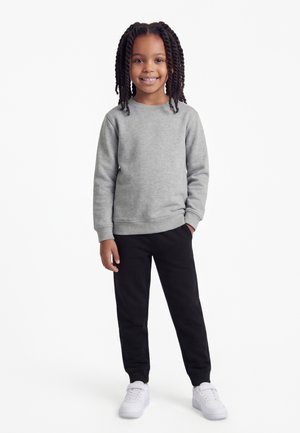 Mosolygó gyerek fonott hajjal, szürke pulóvert, fekete nadrágot és fehér tornacipőt visel, egyik kezét a zsebében tartva áll.