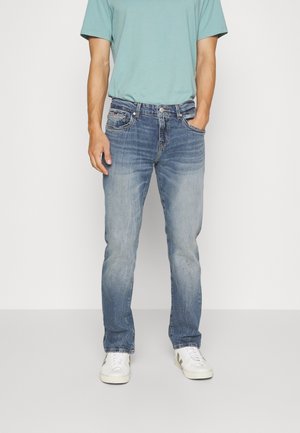 Mężczyzna ubrany w wypłowiałe niebieskie dżinsy, jasnoniebieski t-shirt i białe trampki, stojący z jedną ręką w kieszeni.