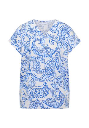 Femme souriante portant une chemise à motifs bleu et blanc, un short en jean, des sandales beiges et un sac à dos foncé, marchant à l'intérieur.