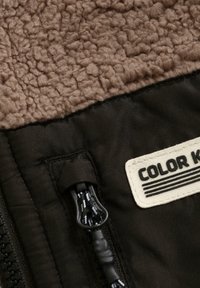 Närbild av en brun fleecejacka som visar en svart dragkedja med snodd och ett rektangulärt patch med texten "COLOR K."