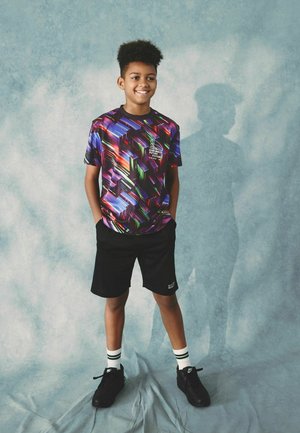 Grafisches T-Shirt in Schwarz, Lila und grünem geometrischen Muster; kombiniert mit schwarzen Shorts und schwarzen Turnschuhen, dazu weiße und grüne gestreifte Socken.