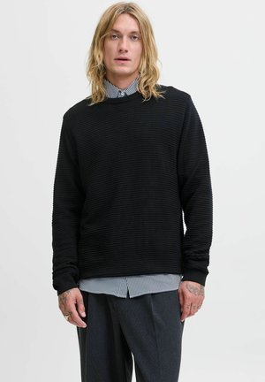 Schwarzer Sweatshirt mit strukturiertem Design, Rundhalsausschnitt und langen Ärmeln, über einem weiß-schwarz gestreiften Hemd getragen, kombiniert mit grauen Hosen.