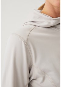Primer plano de una persona con una prenda con capucha de color gris claro que muestra detalle del pespunte en las costuras del hombro y del cuello.