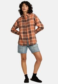 Chemise en flanelle à carreaux dans des tons orange et marron, avec un devant à boutons et des manches longues, associée à un short en denim bleu.