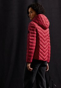 Rode gewatteerde puffer jas met capuchon, met een chevronpatroon en een zijrits. Model draagt donkere jeans en houdt een zwarte riem vast.