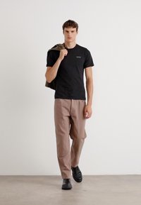 Tricou negru din bumbac cu un logo mic, asortat cu pantaloni bej largi și încălțăminte neagră. Modelul are, de asemenea, o jachetă verde aruncată pe un umăr.