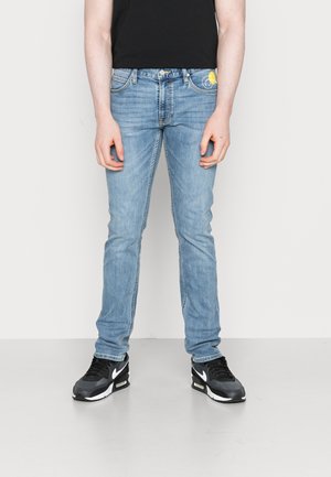 Persona che indossa jeans slim fit azzurri con una piccola grafica gialla sulla tasca, sneaker nere e una maglietta nera.