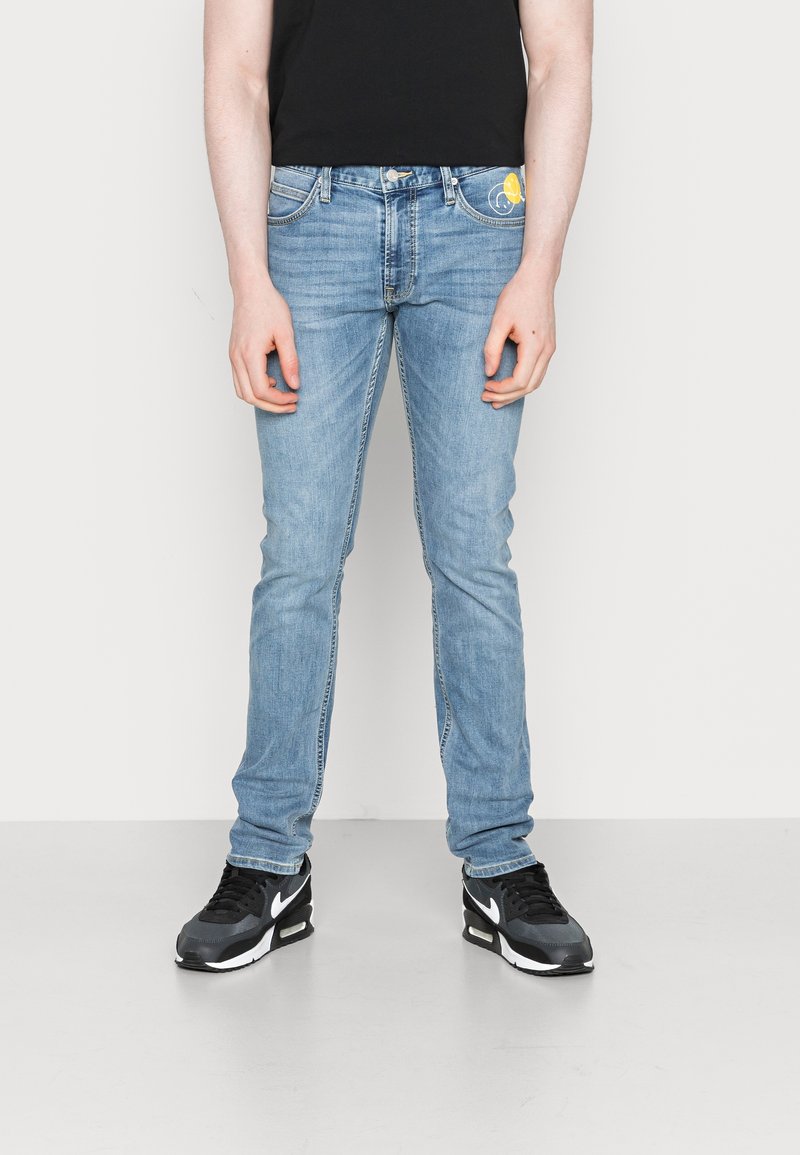 Personne portant un jean slim bleu clair avec un petit motif jaune sur la poche, des baskets noires et un haut noir.