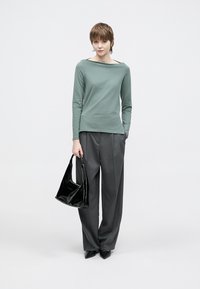 Zelený top s dlhými rukávmi a lodičkovým výstrihom, kombinovaný so sivými širokými nohavicami. Modelka drží čiernu lesklú kabelku a má na nohách špicaté topánky.
