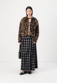 Chaqueta de felpa sintética con cremallera en marrón y negro, combinada con una falda maxi de cuadros en blanco y negro. Zapatos negros sin cordones con tachuelas. Bolso de hombro texturizado.