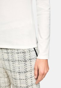 Weißes Langarm-Oberteil kombiniert mit strukturierten Hosen im Karo-Muster in Creme und Schwarz, mit einem dezenten Signaturdetail am Saum.