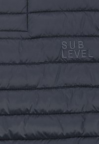 Dunkelblaues, gestepptes Gewebe mit horizontalen Nähten und gesticktem Text "SUB LEVEL" in passendem blauen Faden.