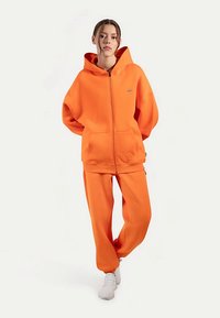 Pomarańczowa bluza na zamek z kapturem i dopasowane spodnie z miękkiego materiału. Bluza posiada duży kaptur i kieszenie z przodu. Dopełnieniem stylizacji są białe trampki.