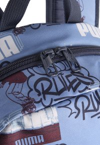 Blå ryggsäck med texturerad tyg, grafiska mönster, svarta dragkedjor och en logotyp. Detaljerade mönster inkluderar stiliserad text och illustrationer.