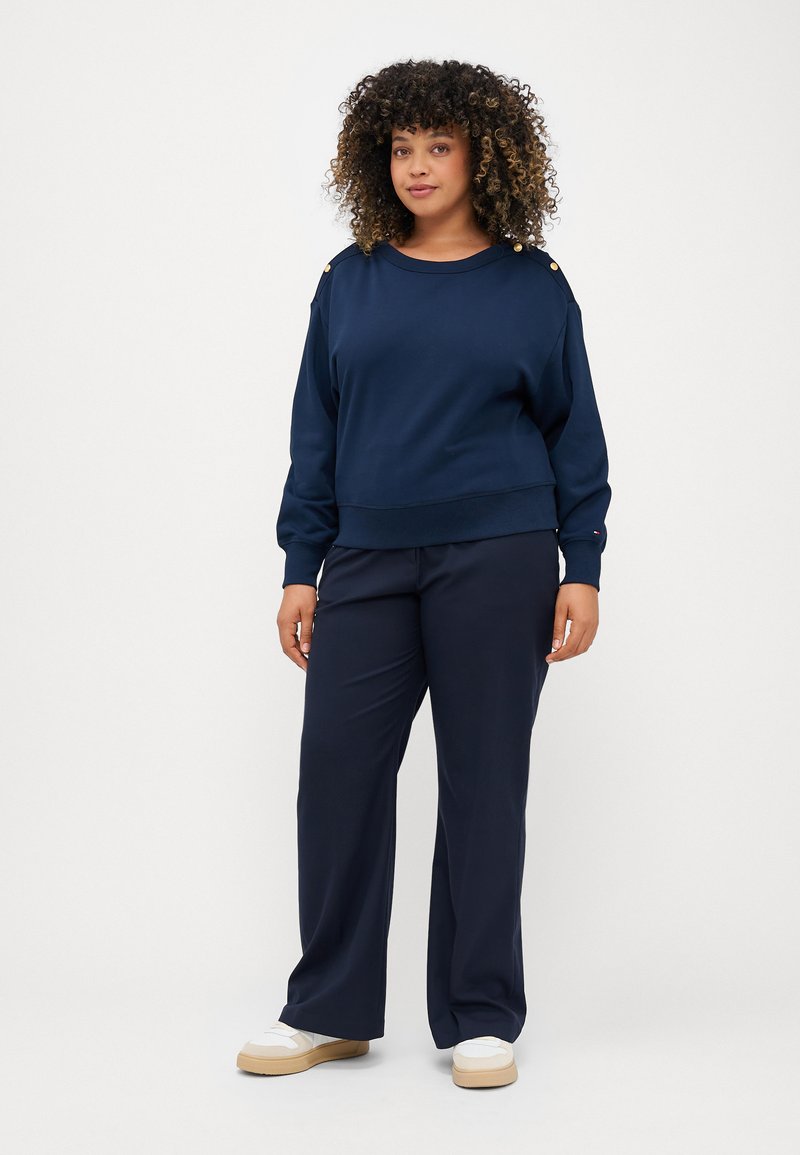 Marineblauwe sweater met geribbelde onderrand en manchetten, voorzien van gouden knopen op de schouders. Gepaard met marineblauwe broek met brede pijpen en lichte schoenen.