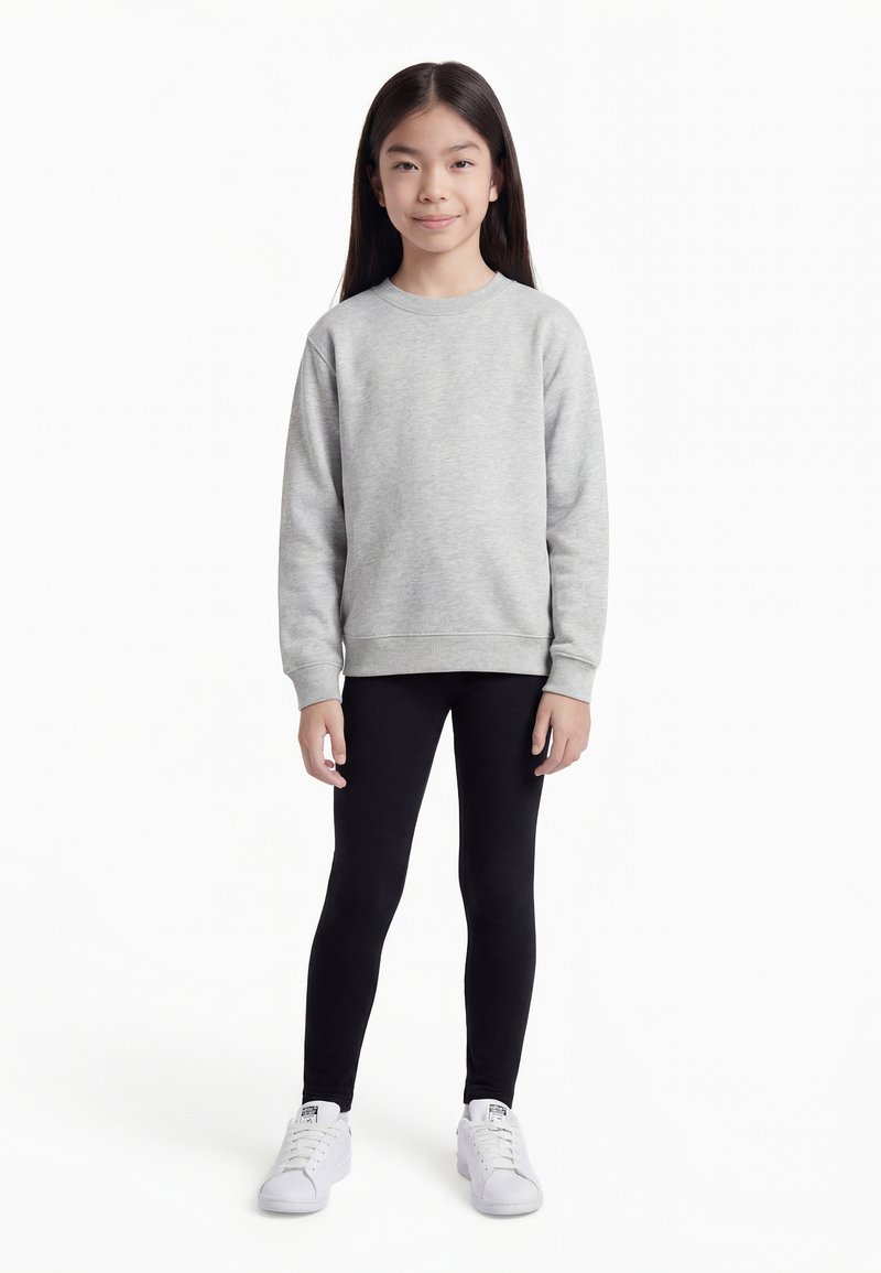 Fiatal lány áll előre nézve, szürke pulóvert, fekete leggingset és fehér sportcipőt visel, fehér háttér előtt.