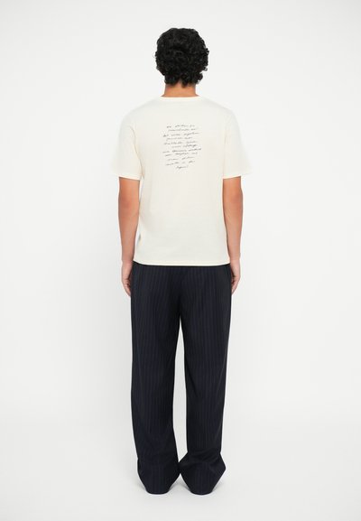 Cremefarvet t-shirt med et håndskrevet tekstdesign på ryggen; parret med løse, mørke stribede bukser.