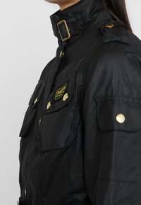 Schwarze Jacke aus glattem Stoff mit hochgeschlossenem Kragen, goldenen Hardware-Elementen, zwei Brusttaschen und Schulterklappen. Barbour-Logo-Patch sichtbar.