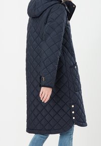 Donna che indossa un lungo cappotto imbottito blu marina con cappuccio, tasche laterali e dettagli in bottoni dorati sopra dei jeans blu, mostrata di lato.