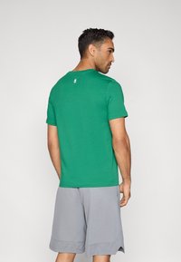 Πράσινο βαμβακερό t-shirt με στρογγυλή λαιμόκοψη, με κοντά μανίκια και λεπτομέρεια λογότυπου στο πίσω μέρος. Συνδυάζεται με γκρι αθλητικές βερμούδες.