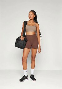 Σπορ εμφάνιση με ένα ταμπά μπραλέτα και καφέ ψηλόμεσα σορτς. Το άτομο κρατά μια μαύρη τσάντα γυμναστηρίου και φοράει μαύρα αθλητικά παπούτσια με λευκές λεπτομέρειες.