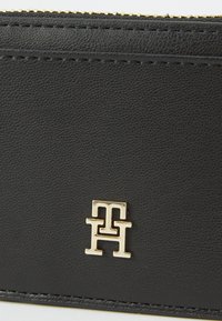Zwarte leren portemonnee met een textuur oppervlakte, voorzien van gouden hardware met een dubbele "T" logo in het midden onderaan. Gouden ritsluiting.