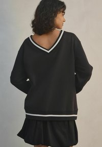 Černá mikina s véčkovým výstřihem, s bílými pruhy na výstřihu a na lemu. Vyrobena z hladkého materiálu, v oversized střihu.