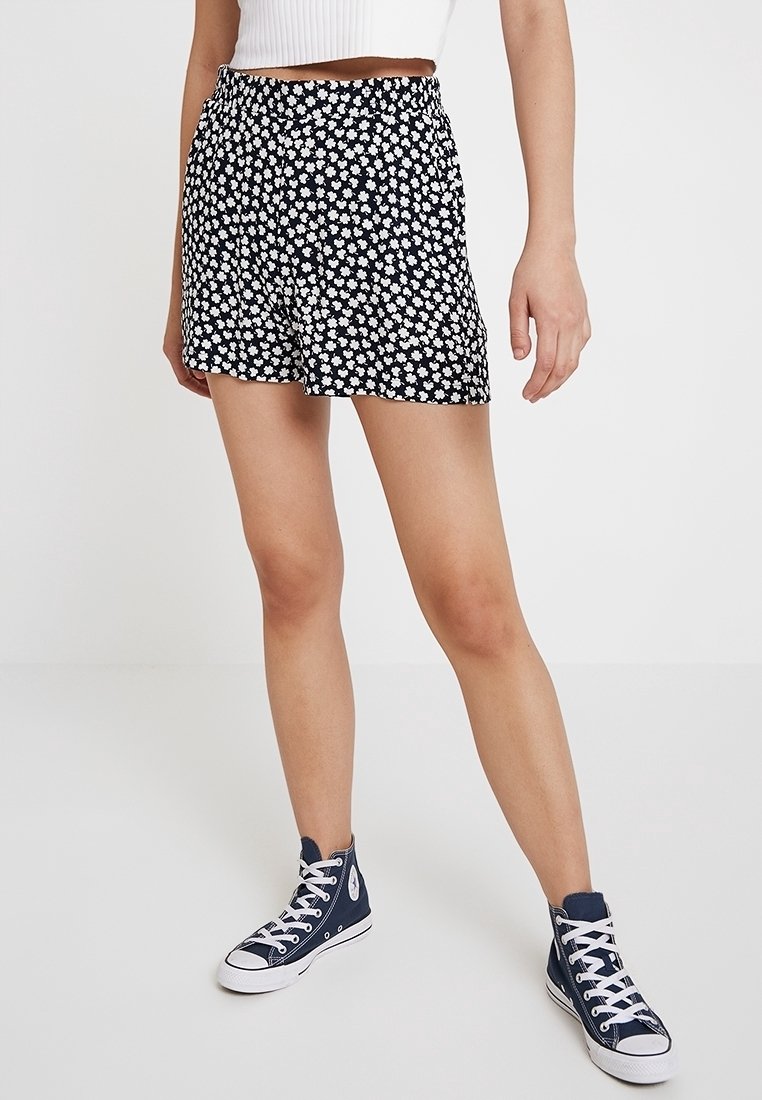 Femme portant un short noir imprimé floral blanc, un top court blanc et des baskets montantes bleu marine, debout sur un sol blanc.