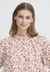 Blouse fleurie avec un fond crème orné de petites fleurs rouges et bleues. Encolure ronde, devant boutonné et manches bouffantes.