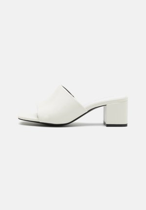 Weißer Mule-Schuh mit offener Spitze, breitem Riemen und niedrigem Blockabsatz auf schlichthintergrund.