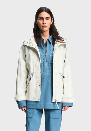 Žena s dugom tamnom kosom, odjevena u plavu majicu na patentni zatvarač i hlače, prekrivena kremastom jaknom s kapuljačom, na jednostavnoj svijetloj pozadini.