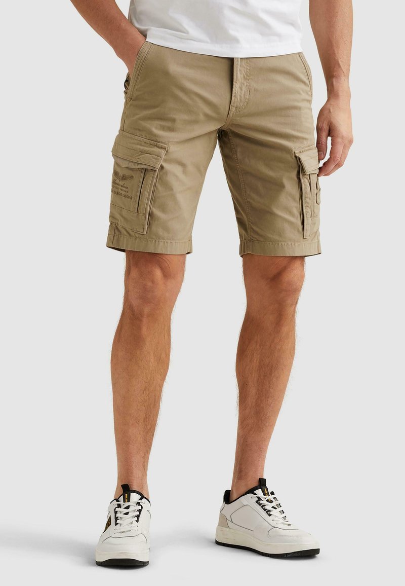 Mann trägt khakifarbene Cargo-Shorts mit Klappentaschen und weißen Sneakers, steht mit den Händen in der Nähe der Taschen vor einem einfarbigen Hintergrund.