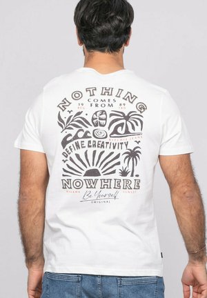 Homme portant un t-shirt blanc avec un motif noir représentant un soleil, des palmiers, des vagues, et le texte "Rien ne vient de nulle part, définissez la créativité, Soyez vous-même" au dos.