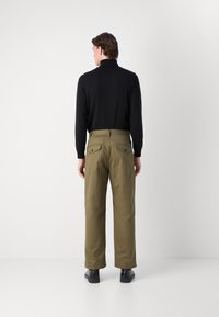 Pantalon en coton vert olive avec une coupe décontractée, jambes droites et deux poches arrière. Porté avec un haut noir à manches longues et des chaussures noires.