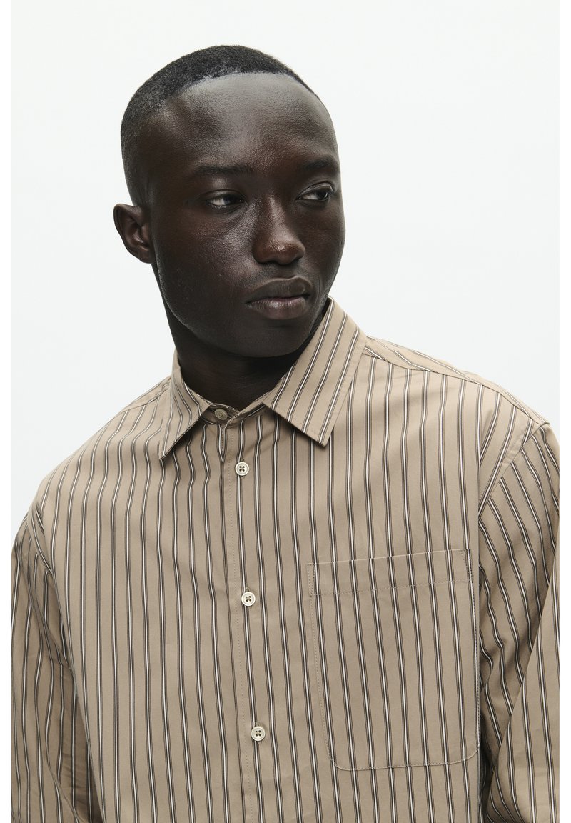 Camicia beige con bottoni, a righe verticali sottili in tonalità scure, colletto a punta, taschino sul petto e texture liscia.